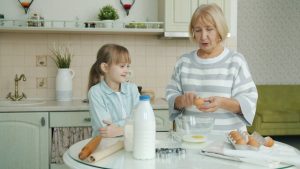 Senior woman is cooking pastry in kitchen teaching little girl discussing ingredients and breaking eggs in bowl. Cookery, family and people concept.